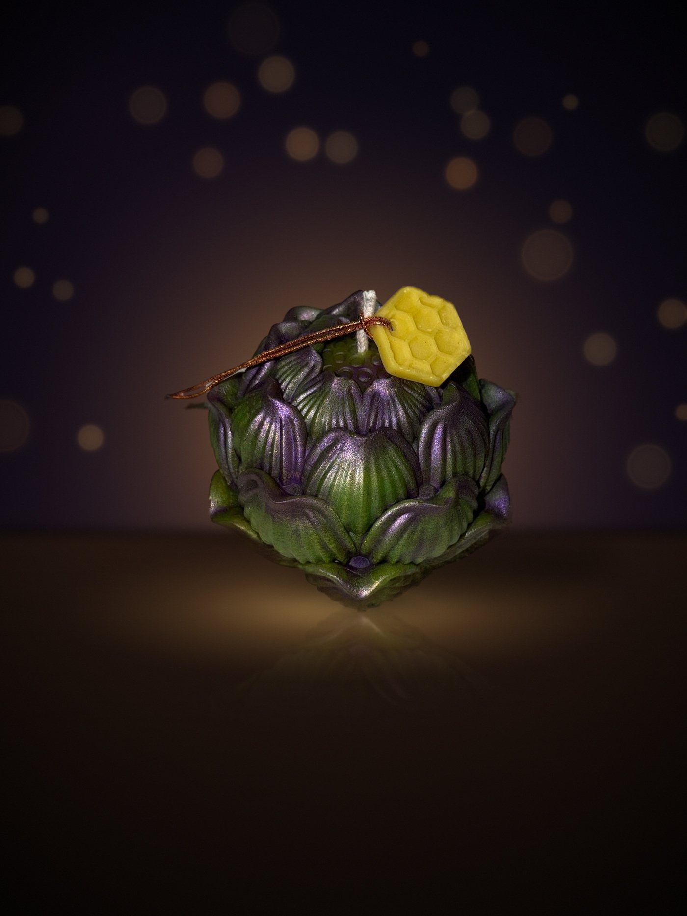 Maine beeswax artichoke candle, three-quarter view, forest green with purple mica and honeycomb tag, warm candlelit background