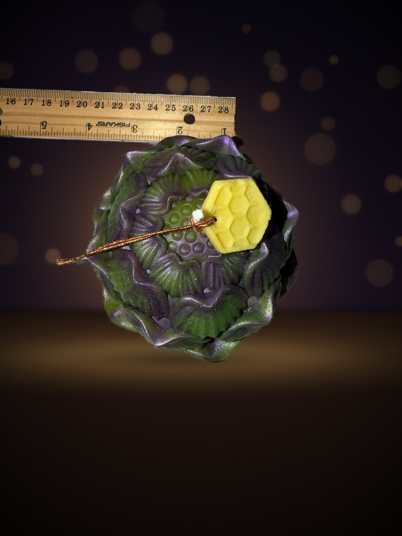 Maine beeswax artichoke candle seen from above next to a ruler for size, honeycomb tag, warm candlelit background