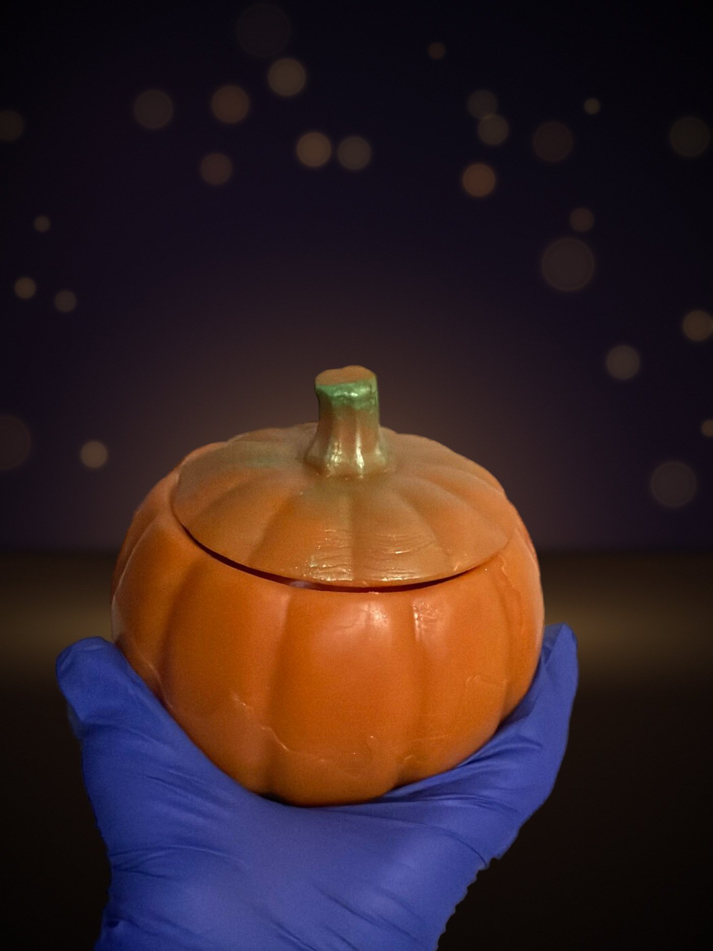 Maine beeswax pumpkin candle, jack-o’-lantern shape, held in a gloved hand, warm candlelit background