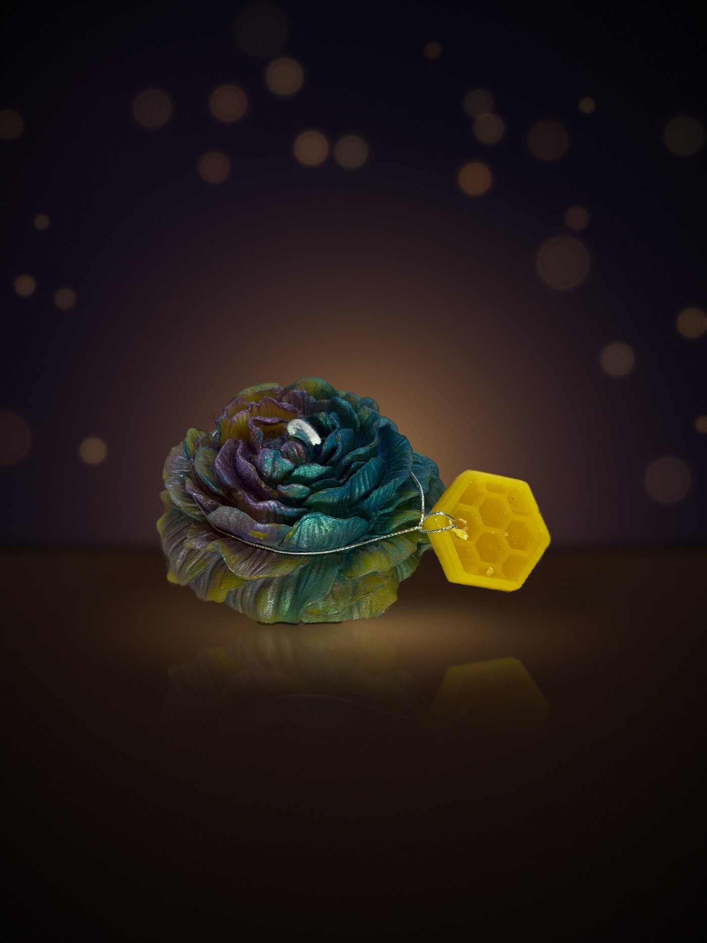 Maine beeswax rose candle in teal, purple and gold with honeycomb tag, warm candlelit background